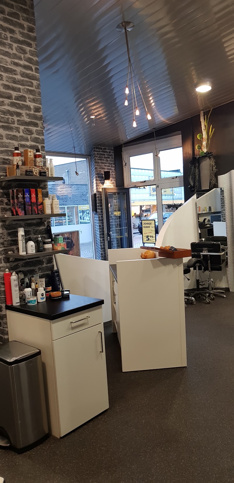 De voorkant van de salon met bakstenen muur, hanglampen en producten op de planken; door het raam is de Jumbo te zien.